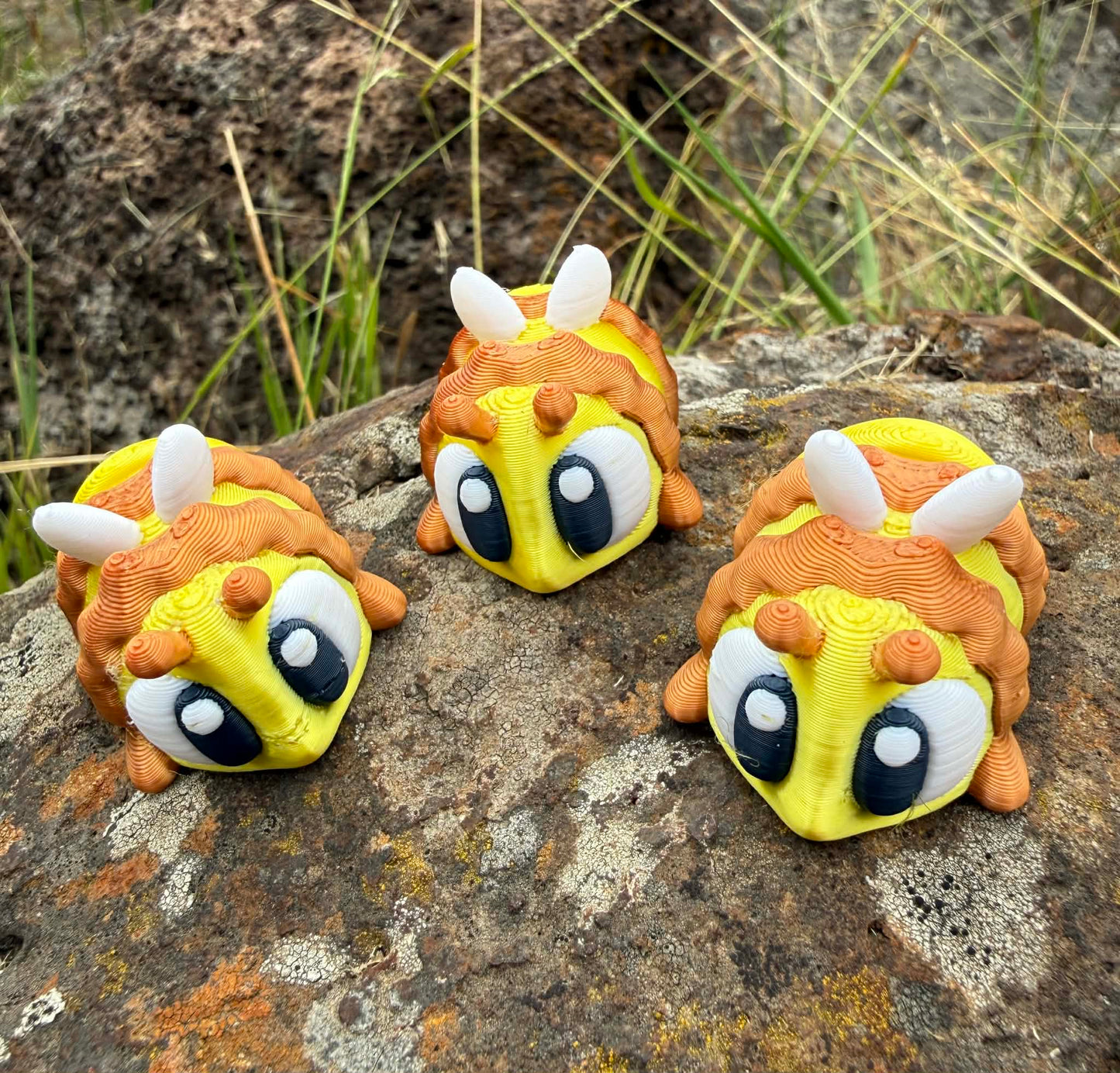 Three cartoonish 3D printed bee-like creatures on a rocky surface with grass in the background