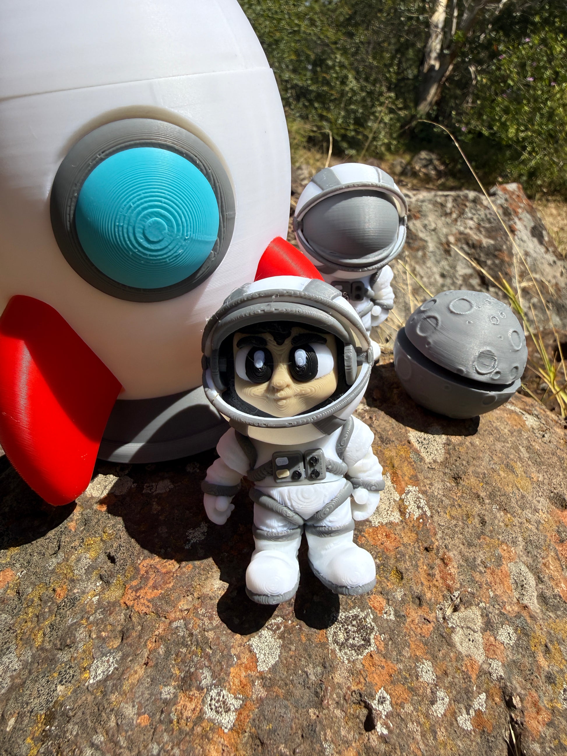 3d printed rocket with a white body, red accents, and gray and blue details on a rocky surface. with two astronauts and moon clicker
