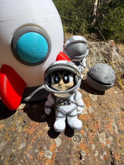 3d printed rocket with a white body, red accents, and gray and blue details on a rocky surface. with two astronauts and moon clicker