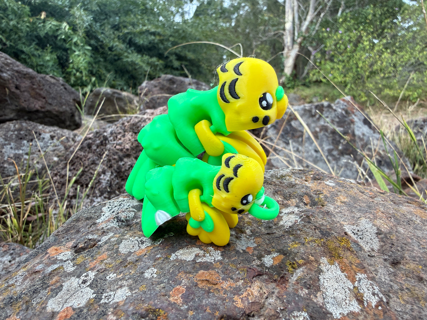 Two green and yellow toy 3d printed budgies. one keyring size on a rock with a natural background