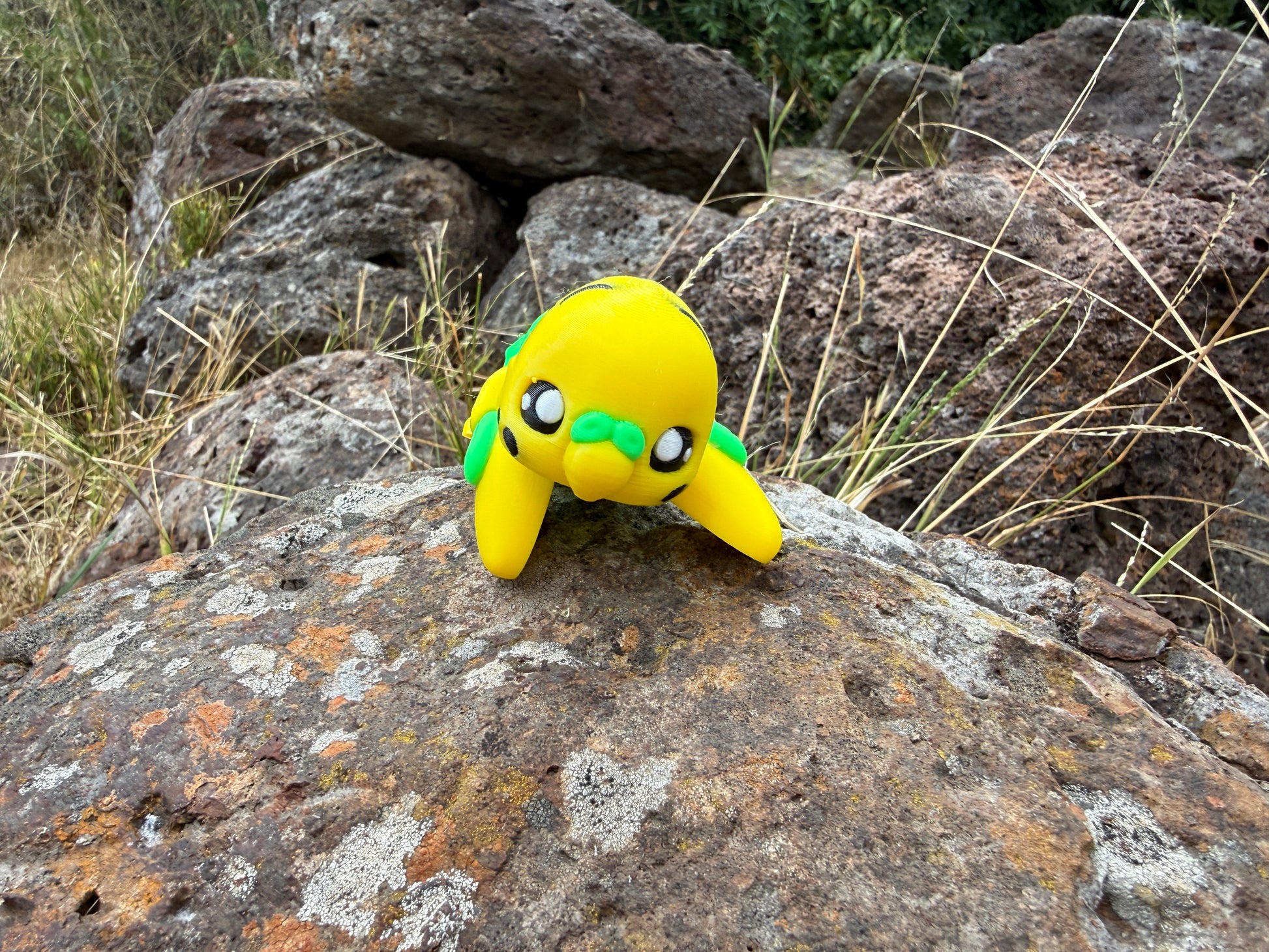 Yellow 3d printed budgie with green accents on a rock in a natural setting
