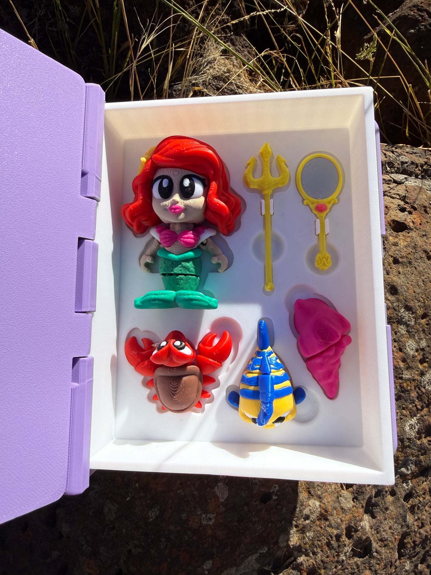 Set of colorful 3d printed articulated figurines (mermaid, yellow and blue fish, crab, cone shell, trident, mirror) in a white display case (book open) with a purple base, against a natural background.