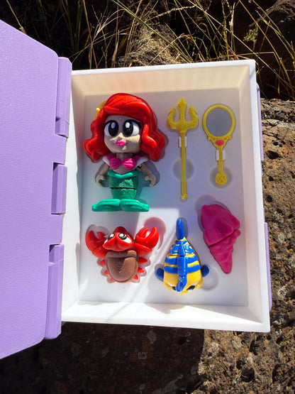 Set of colorful 3d printed articulated figurines (mermaid, yellow and blue fish, crab, cone shell, trident, mirror) in a white display case (book open) with a purple base, against a natural background.