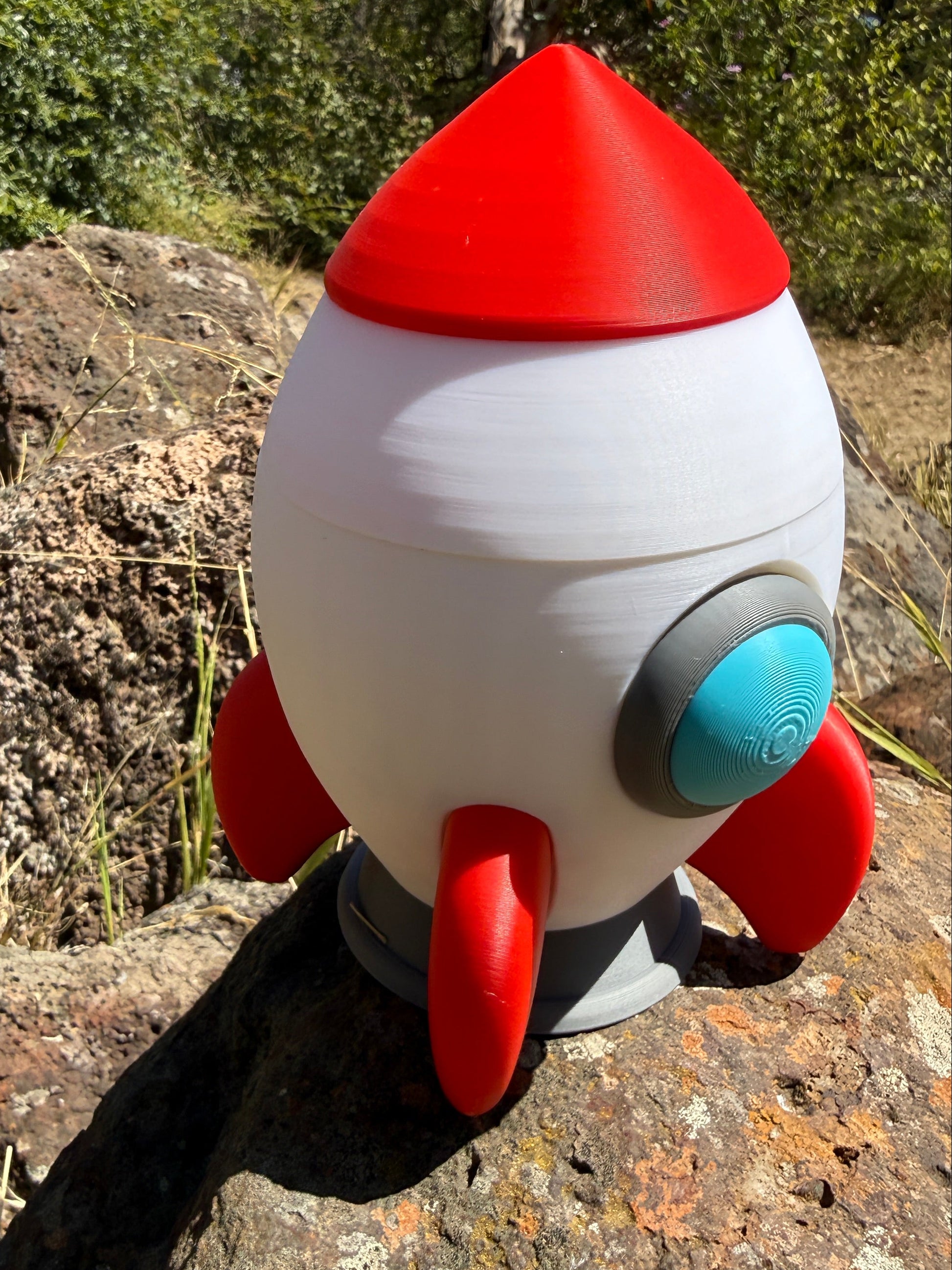 3d printed Toy rocket that is red, white, grey and light blue window on a rocky surface with greenery in the background