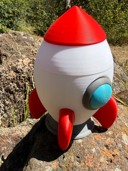 3d printed Toy rocket that is red, white, grey and light blue window on a rocky surface with greenery in the background
