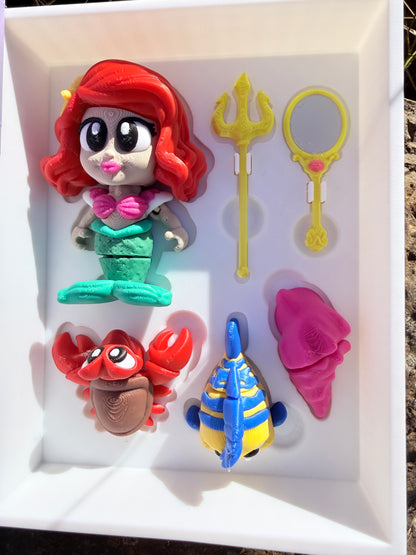 Set of colorful 3d printed articulated figurines (mermaid, yellow and blue fish, crab, cone shell, trident, mirror) in a white display case (book open) with a purple base, against a natural background.