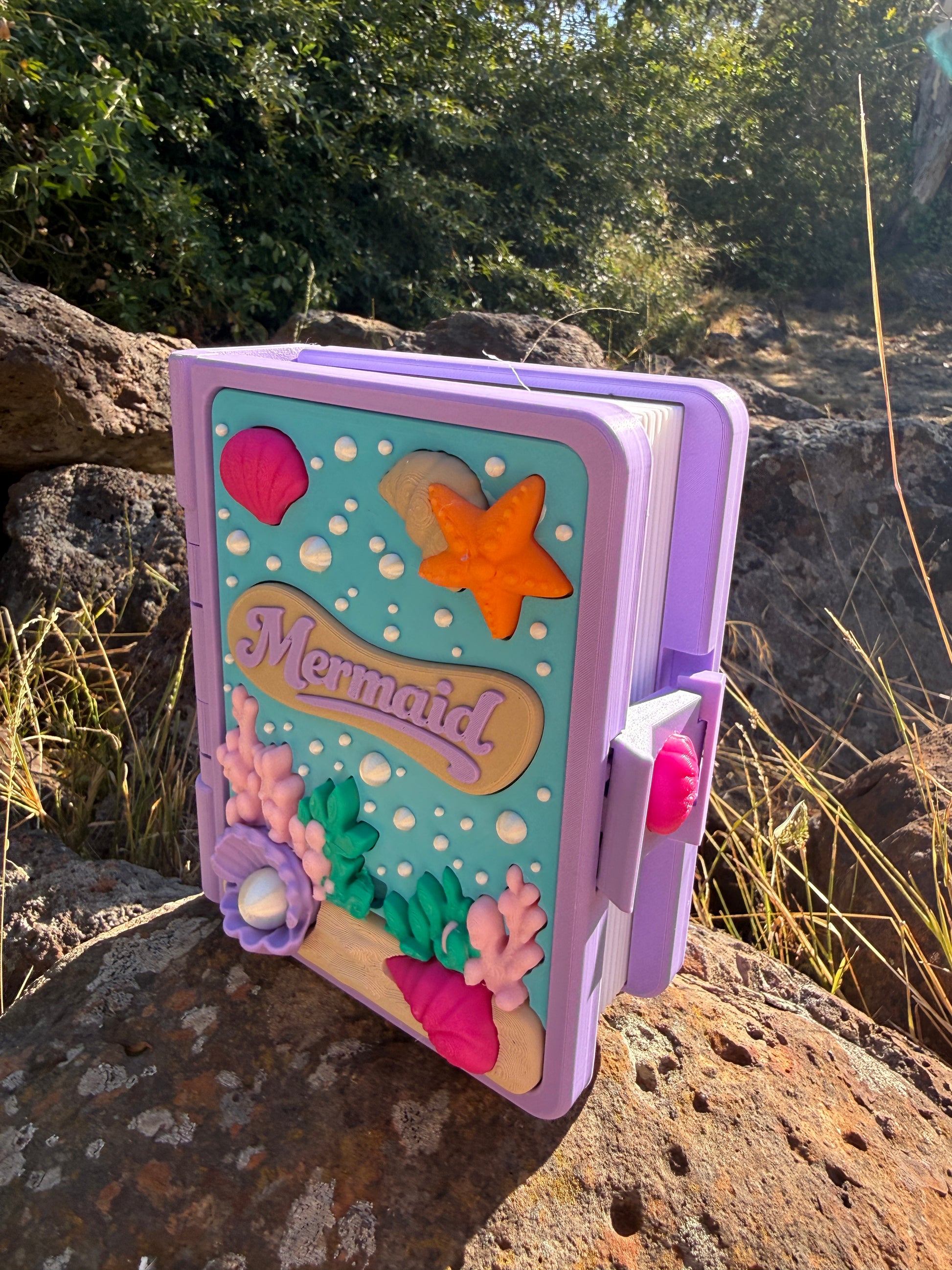 Colour purple, blue, white, green, pink 3d printed book with mermaid design on a rock