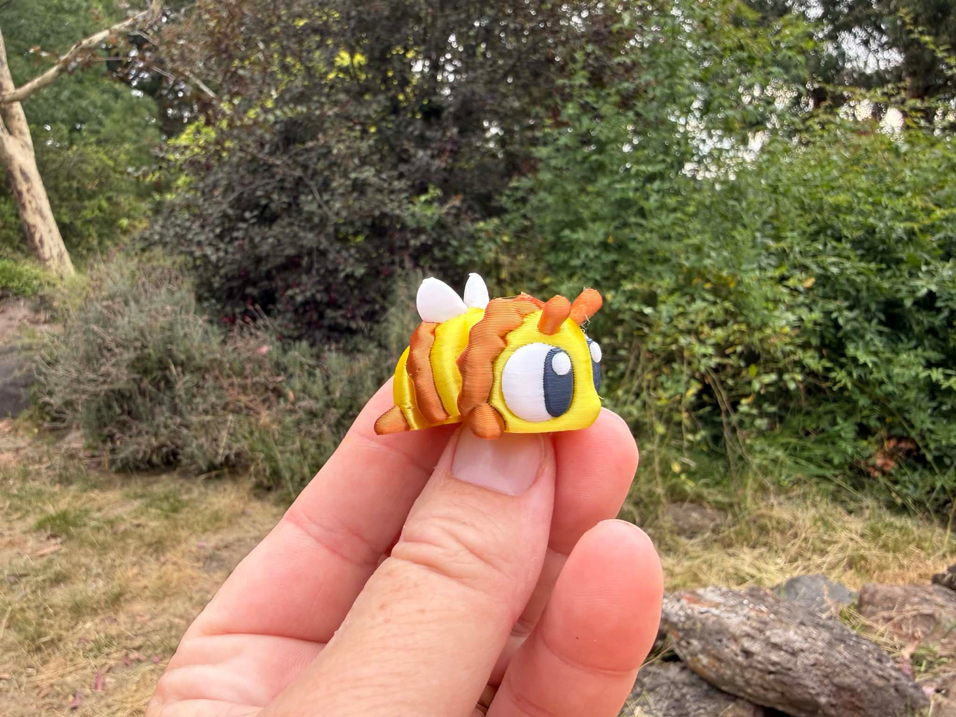 Hand holding a small yellow and brown 3D printed bee with googly eyes against a natural background