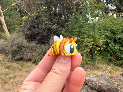 Hand holding a small yellow and brown 3D printed bee with googly eyes against a natural background