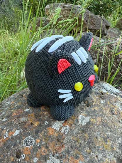 Knitted cat money box 3d printed black and white on a rock with grass in the background