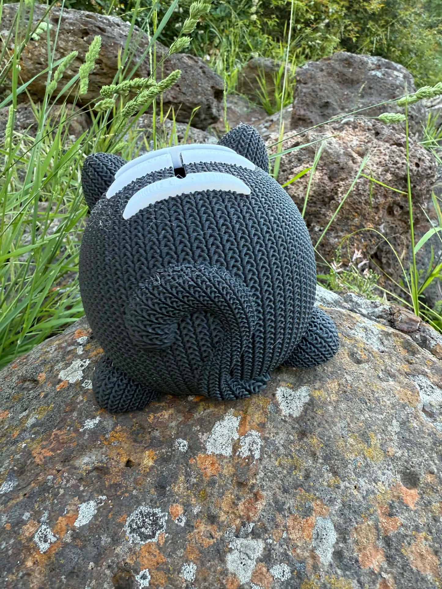 Knitted cat 3d printed money box on a rock with grass and rocks in the background