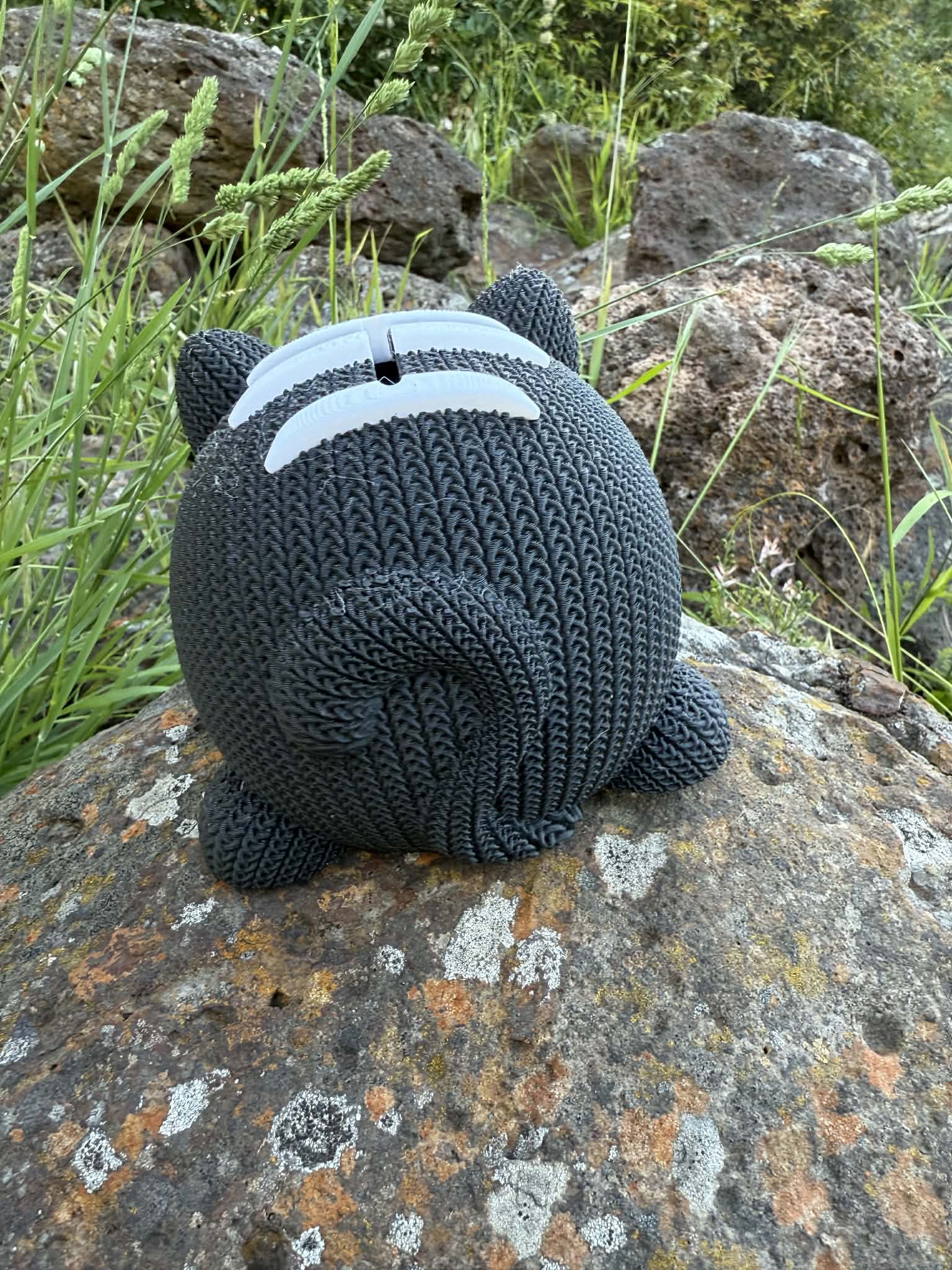 Knitted cat 3d printed money box on a rock with grass and rocks in the background