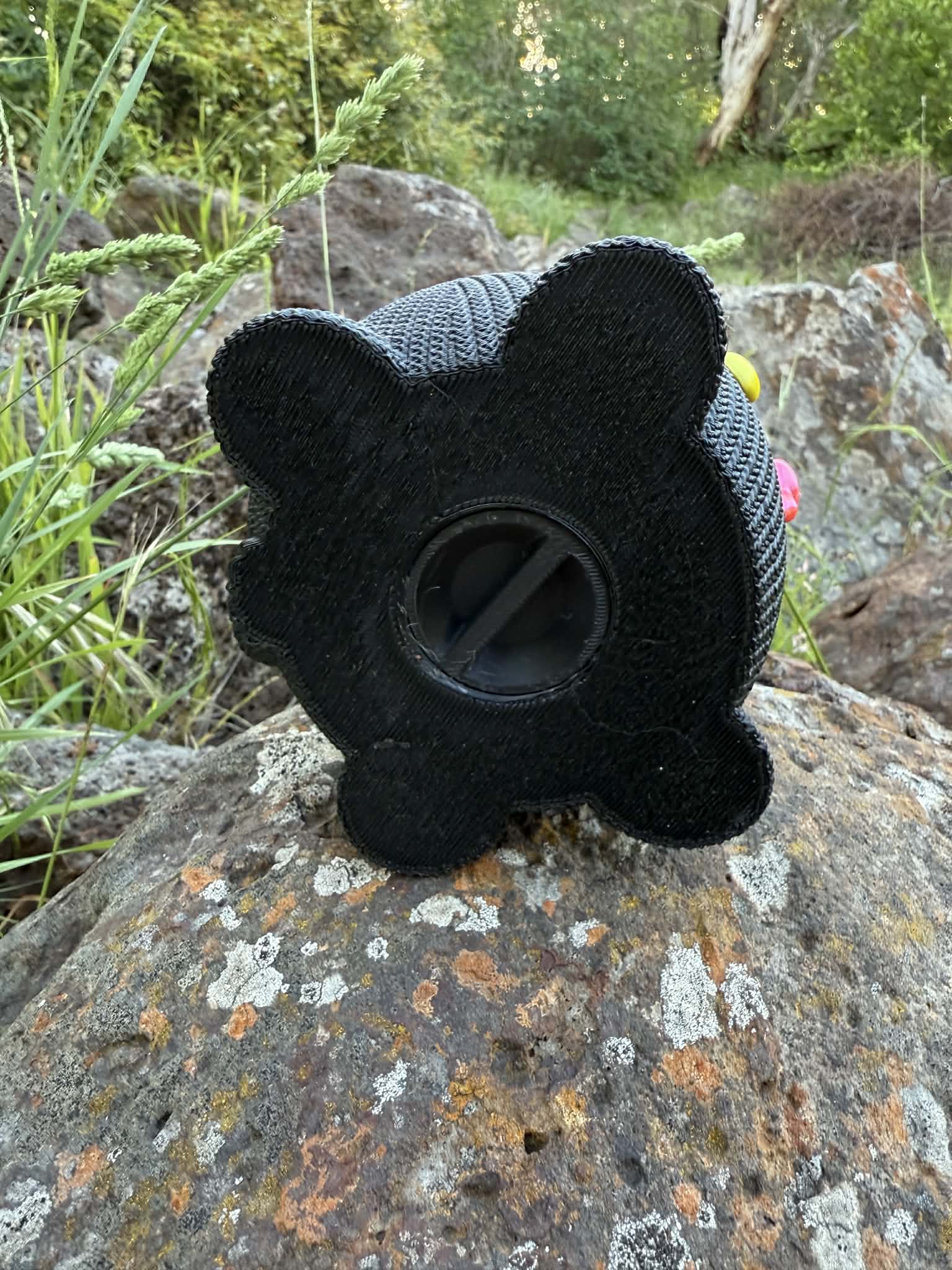 Black knitted cat-shaped money box 3d printed showing screw in bottom on a rock with greenery in the background