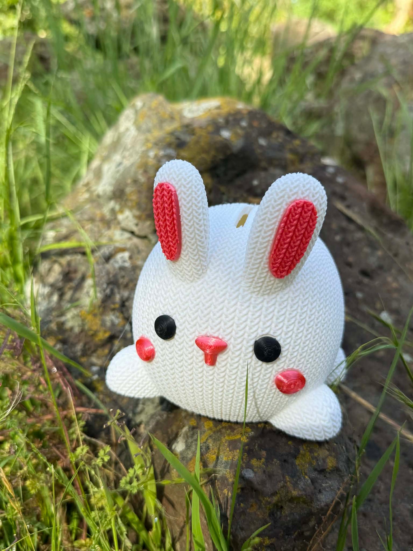3D printed White knitted bunny moneybox with pink accents on a rock in a natural setting
