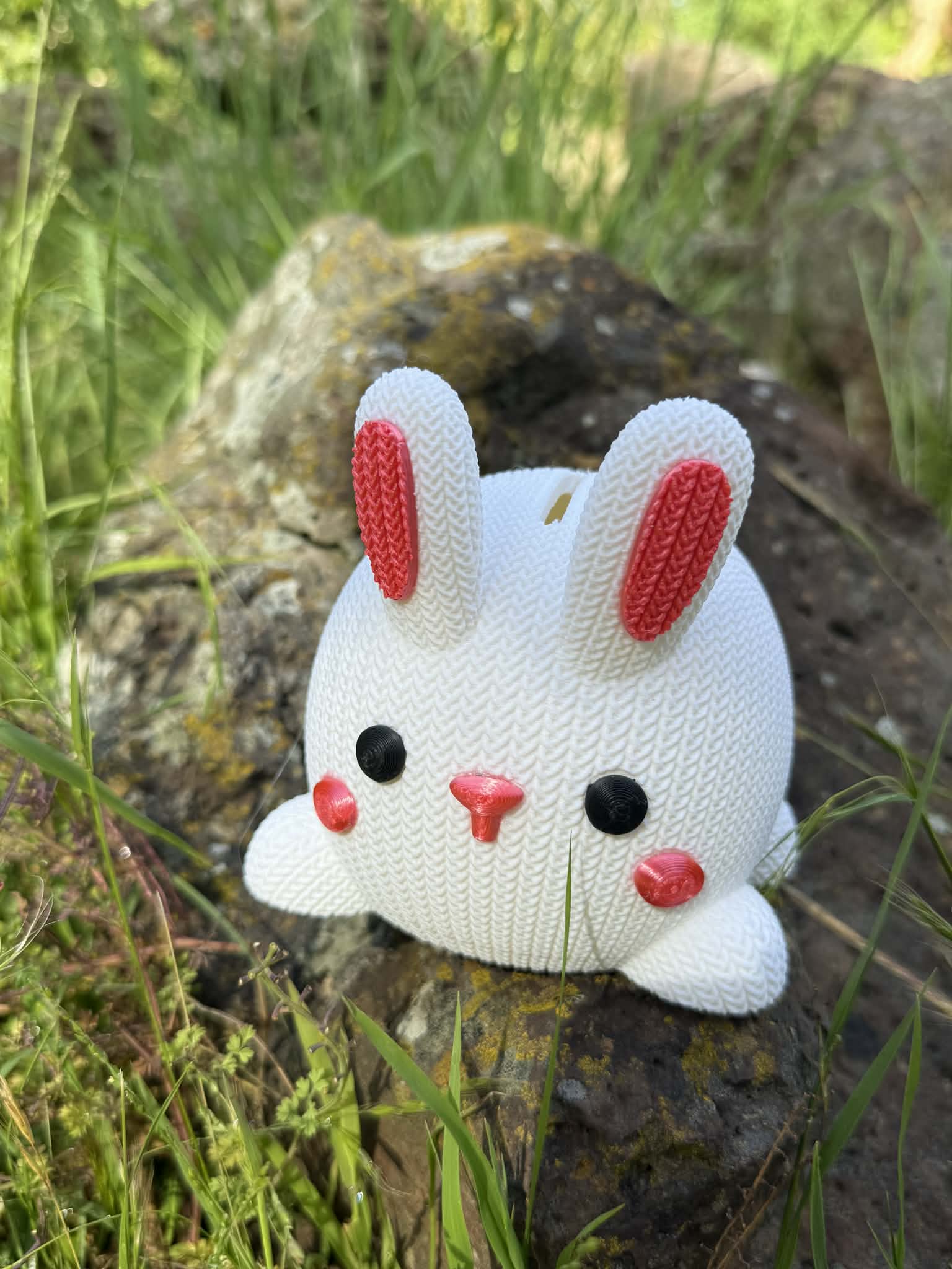 3D printed White knitted bunny moneybox with pink accents on a rock in a natural setting