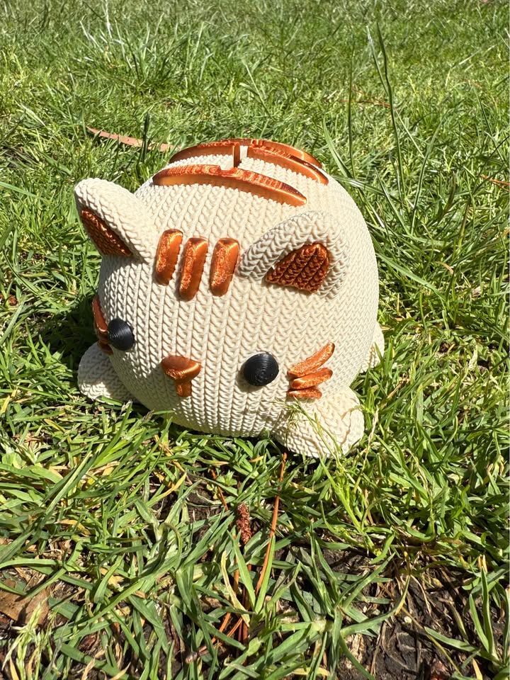 Beige and copper Knitted cat-shaped 3D printed money box on grass