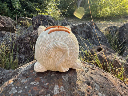 Knitted back of cat money box showing tail. beige and copper colour  on a rock with natural background
