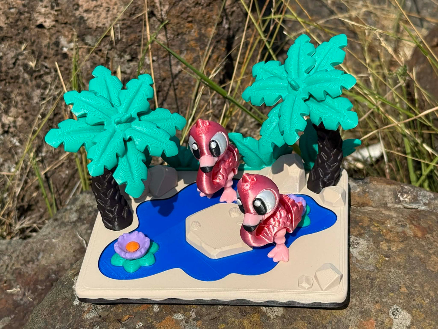 Two pink flamingoes 3d printed articulated characters on a miniature landscape with palm trees and a pond.