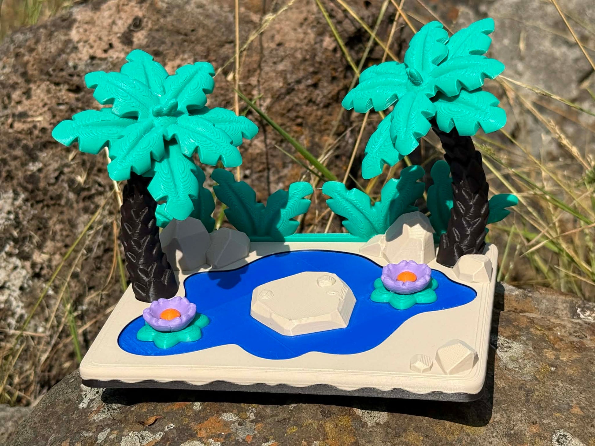 3d printed landscape with palm trees, a pond, and flowers on a rocky surface 