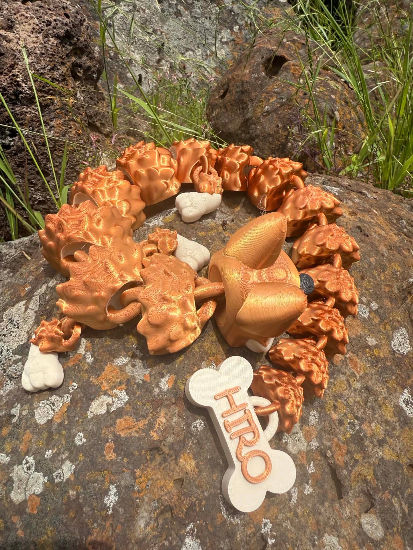 3D printed German Shepard dragon coiled around on a rocky surface with a 'HIRO' bone shaped tail tag.