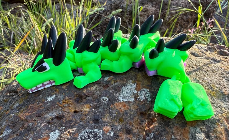 Green 3d printed mini dragon with black horns and two fists for a tail end on a stone surface