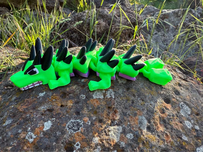 Green mini dragon 3d printed on a rocky surface with grass in the background