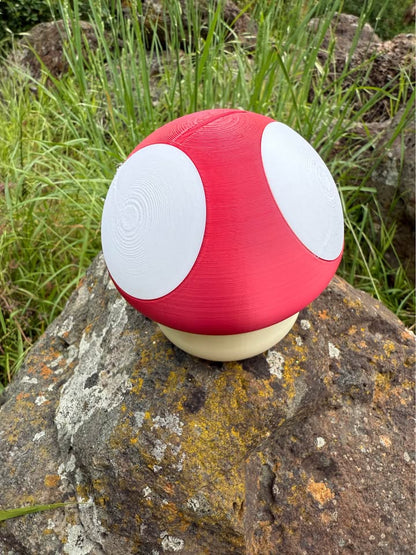 Red and white mushroom-shaped 3d printed money box on a rock with grass in the background