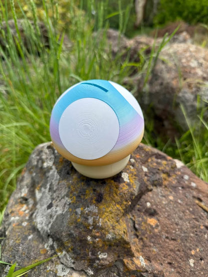 3d printed money box with yellow, pink and blue top. with white spots. and beige bottom on a rock with grass in the background