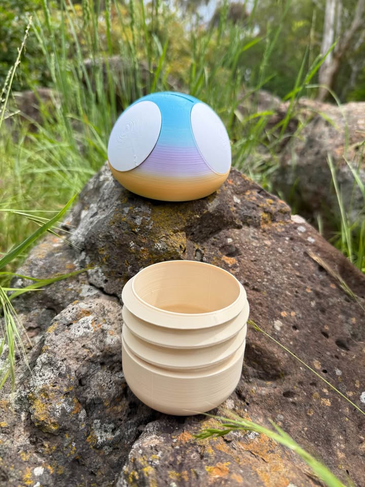 3d printed top of money box gradient-colored and the bottom one beige, on a rocky surface with grass in the background.