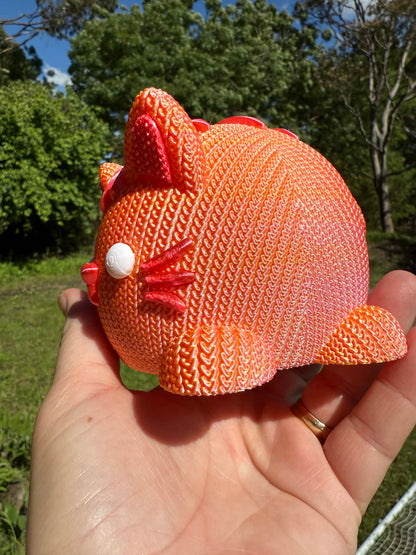 Hand holding a knitted orange cat money box 3d printed with a blurred natural background