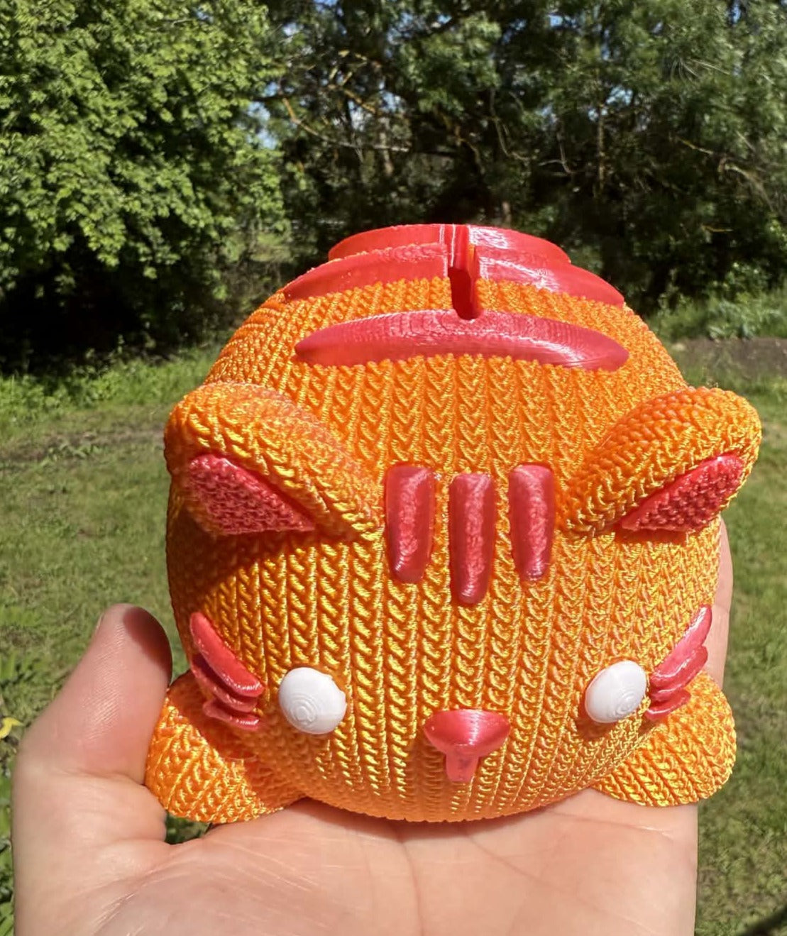 Hand holding an orange and pink knitted cat-shaped money box 3d printed against a natural background