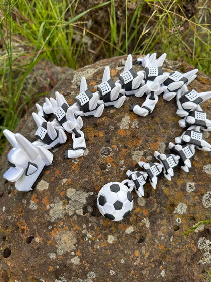 White and black 3d printed dragon with a soccer ball tail and a white hat with text. arranged in a circle on a textured stone surface with grass in the background.