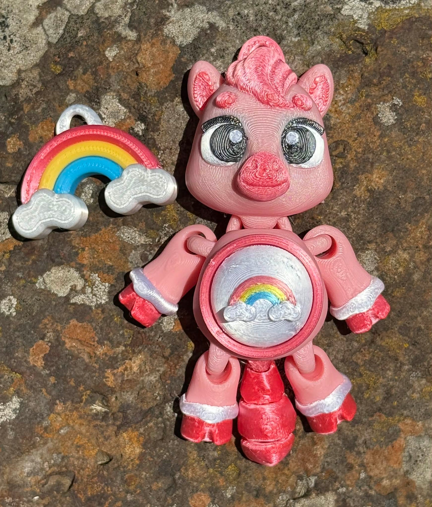 Pink pony 3D printed with rainbow design on the tummy with a rainbow next to it on a textured surface