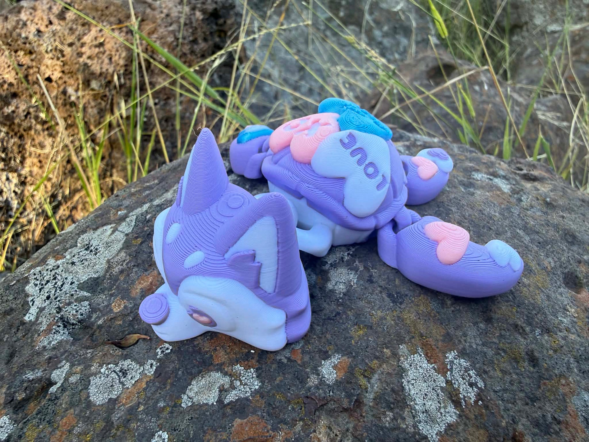 Pink and purple 3d printed turtle/husky on a rock with grass in the background
